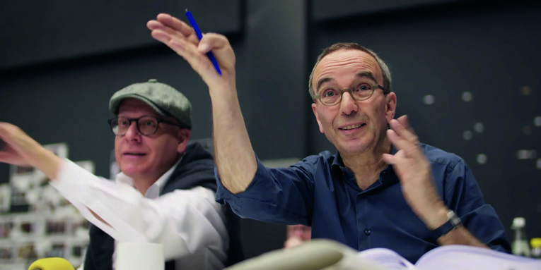 Auf dem Bild sind zwei Personen zu sehen: links Chefdramaturg Sergio Morabito mit Schirmmütze auf dem Kopf und rechts Intendant Jossi Wieler. Die beiden sitzen nebeneinander an einem Tisch und geben anderen Personen Handzeichen.